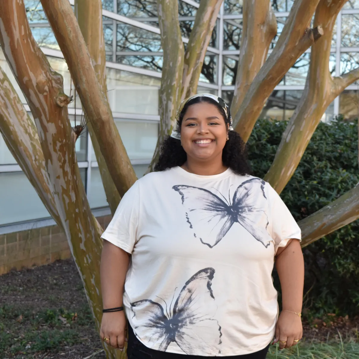 person standing in a butterfly t-shirt smiling with white bandana in front of tree
